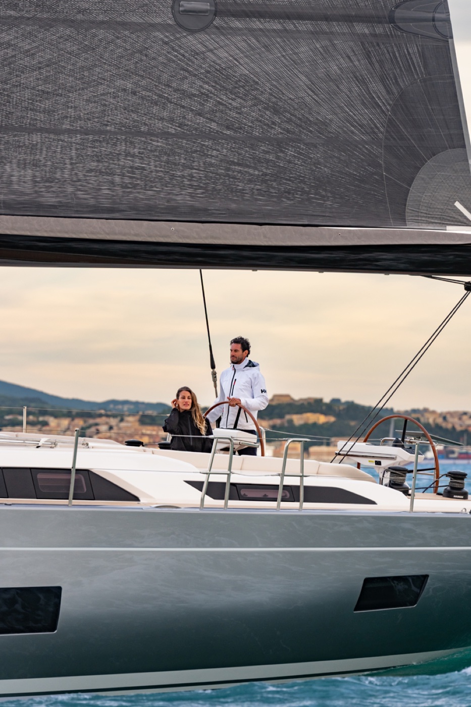 Um casal ao timão do Grand Soleil 60 durante a navegação, com o litoral ao fundo. Foto: Gianluca Napthalina Camporesi / Grand Soleil Yachts