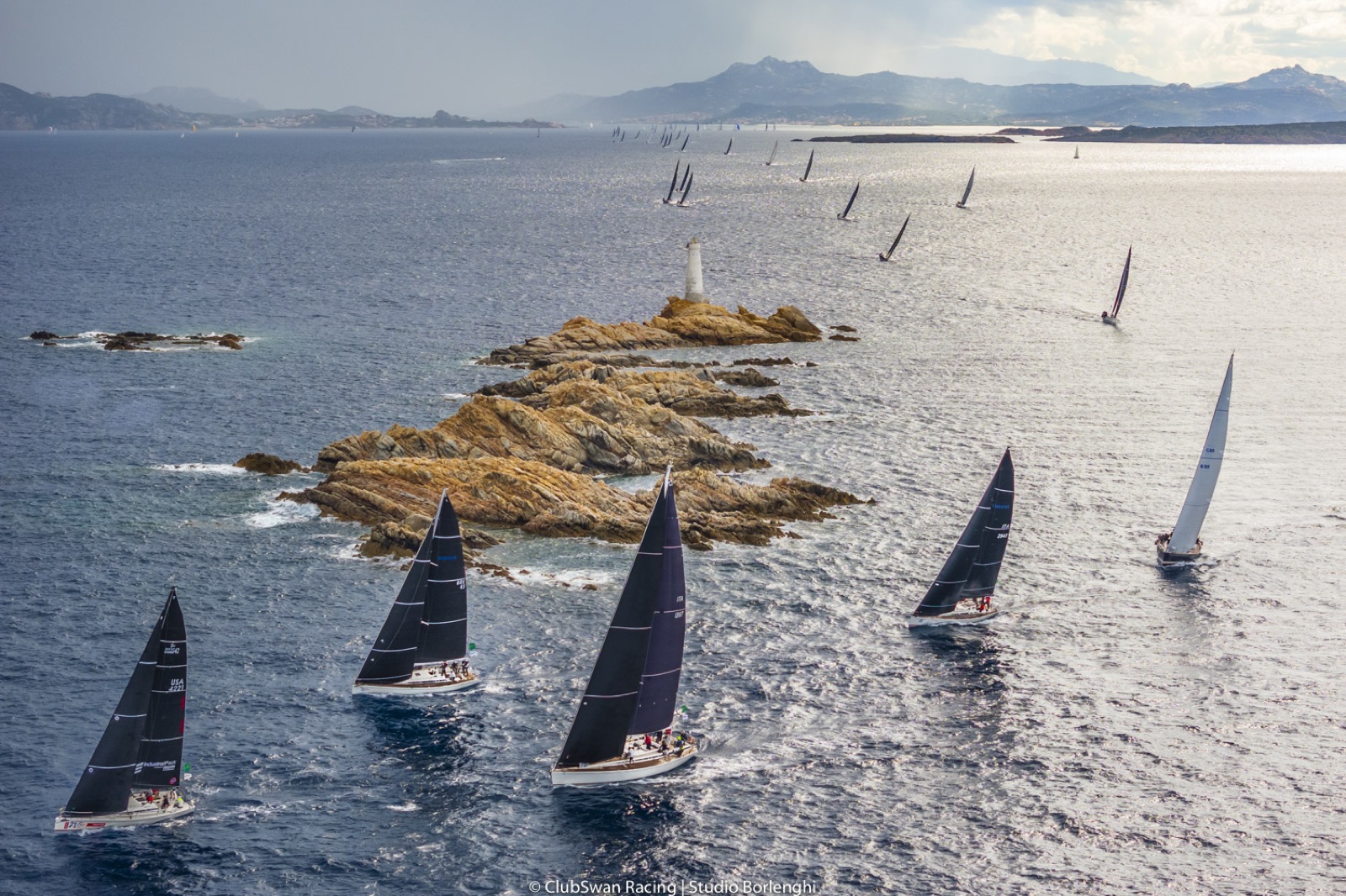 A frota de veleiros Swan em regata, com a costa montanhosa da Sardenha ao fundo. Foto: Studio Borlenghi / ClubSwan Racing